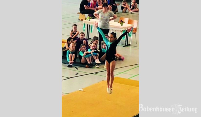 Dilara beim Strecksprung am Boden, im Hintergrund das Team.